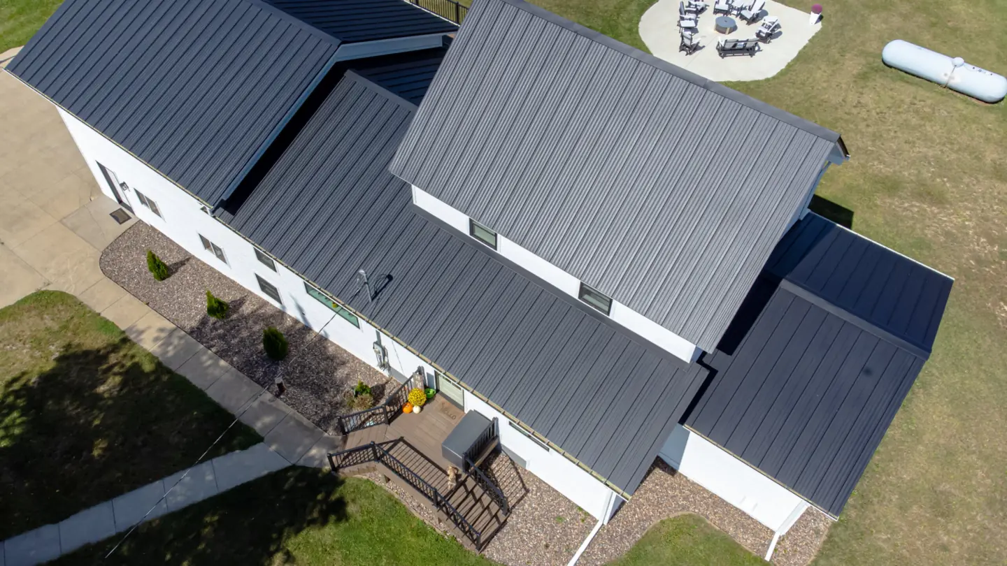 Overhead drone view of a white two-story home with a dark standing-seam metal roof, a back deck, gravel landscaping and a round fire-pit patio in a large lawn.