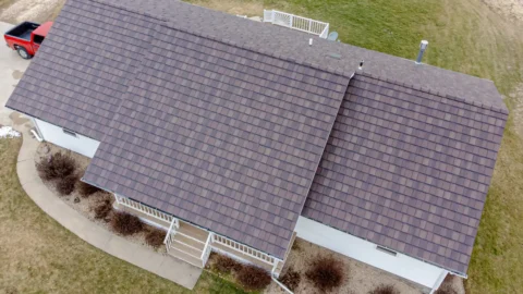 Angled drone view of a ranch-style house with a brown shingle roof, white siding and a front porch railing, with a red pickup truck parked in the driveway.