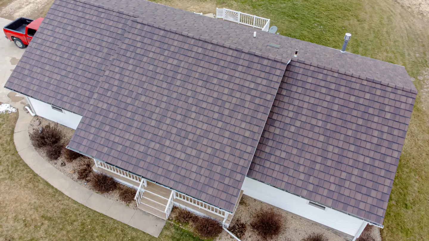 Angled drone view of a ranch-style house with a brown shingle roof, white siding and a front porch railing, with a red pickup truck parked in the driveway.